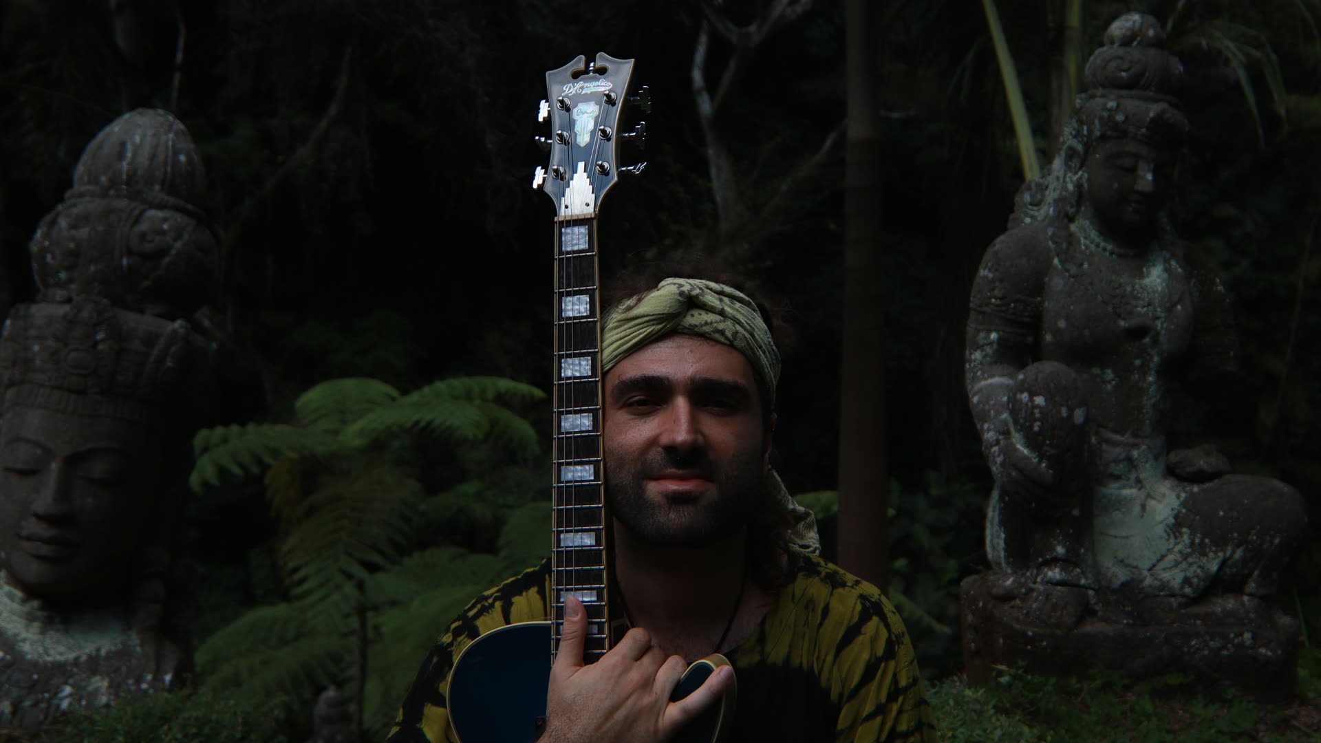 Constantine Cheimarios holding a D'Angelico archtop guitar in a tropical garden of stone statues