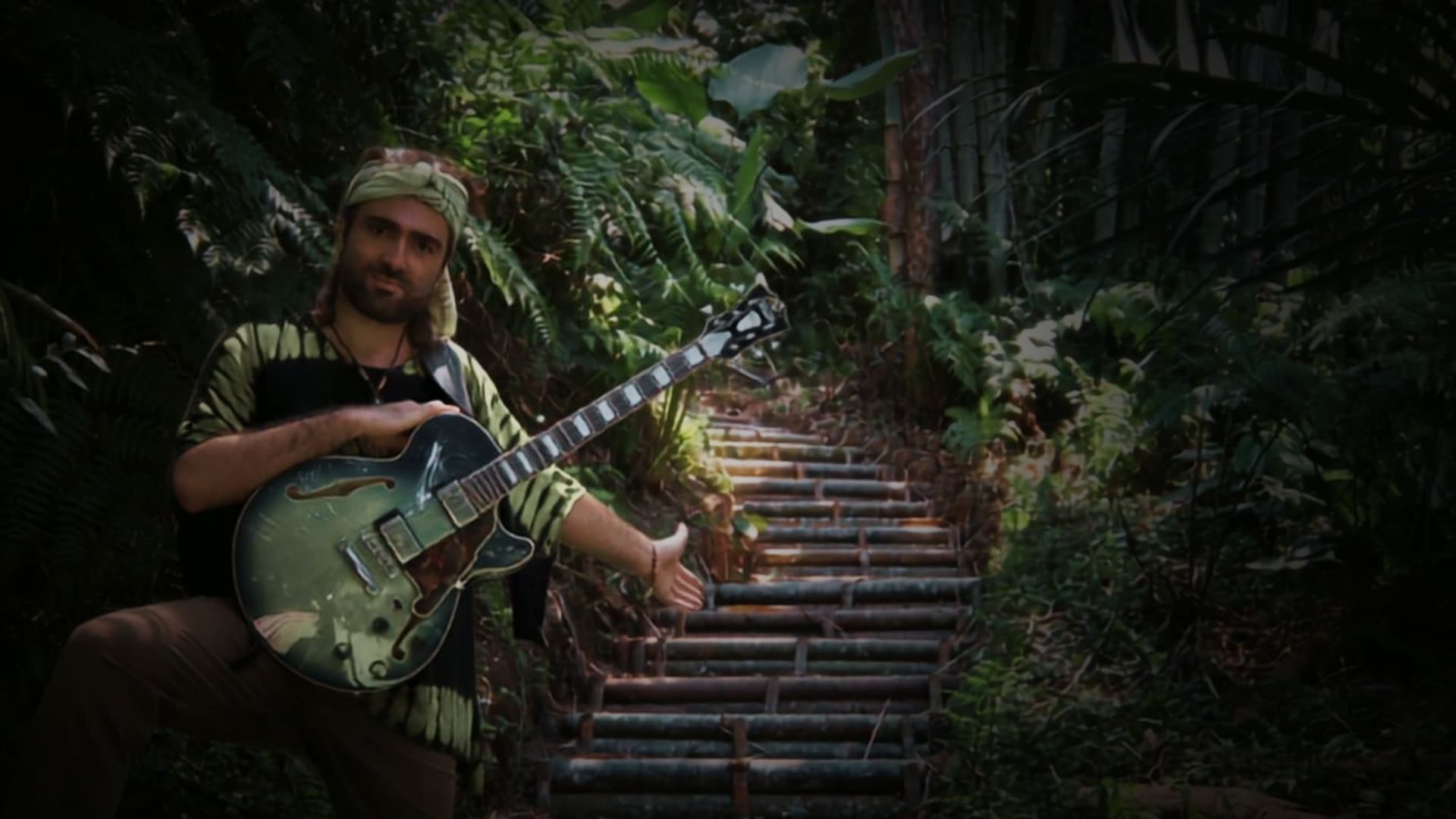 Constantine seated with his archtop guitar at the foot of a staircase climbing through the jungle